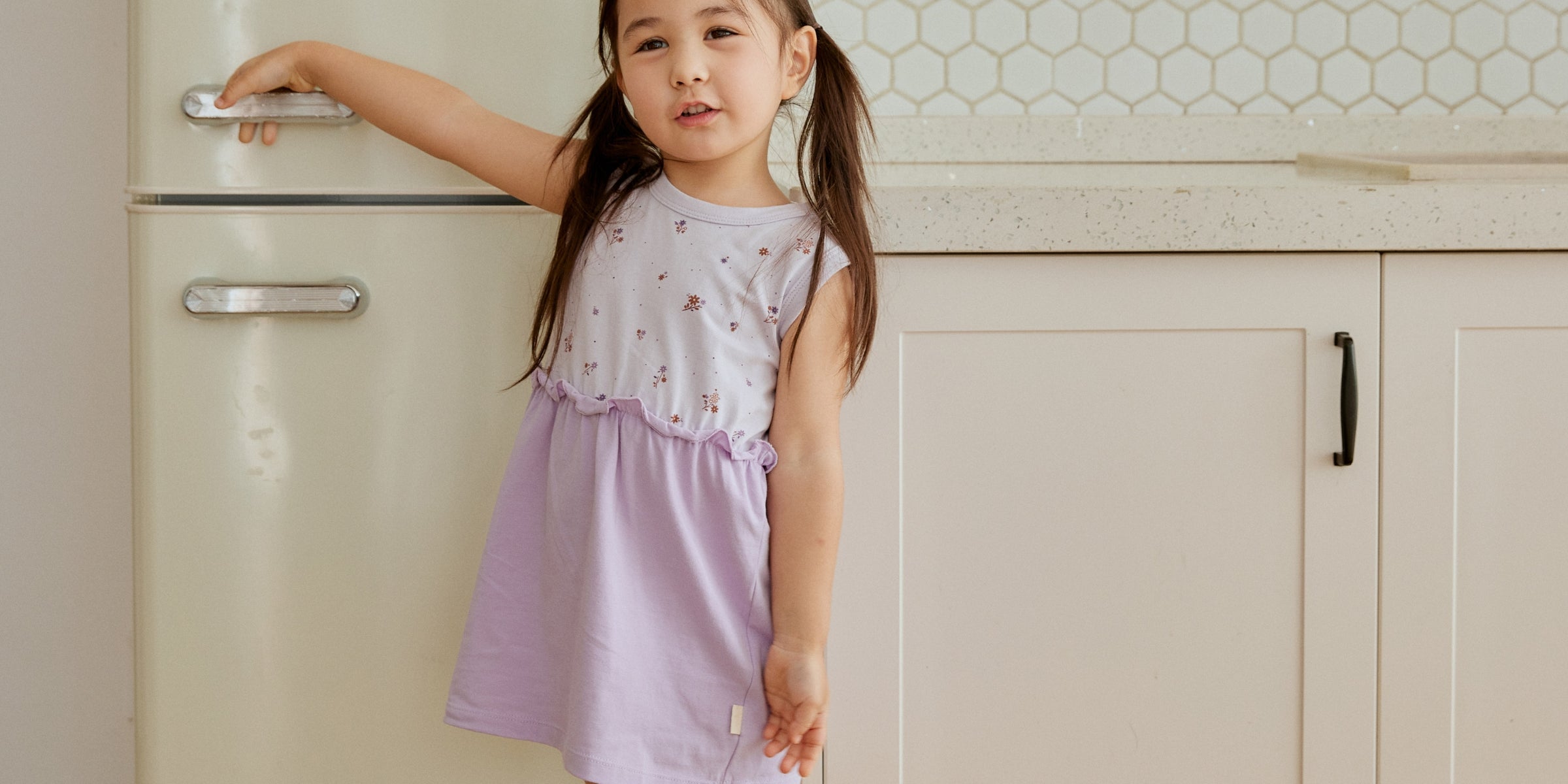 toddler girl wearing a purple dress and standing beside the fridge