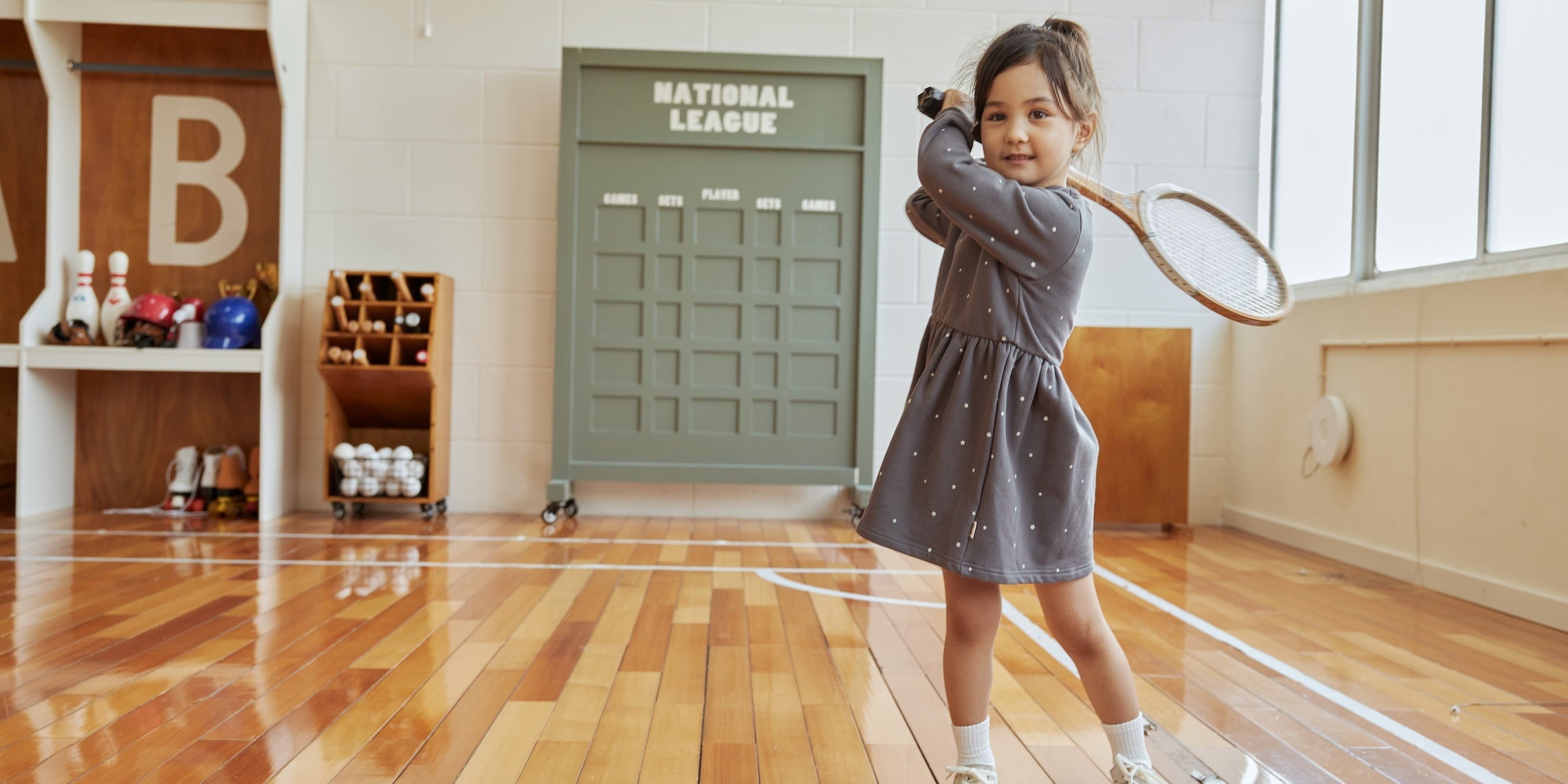 Child in a gray dress holding a badminton racket in an indoor setting with wooden floor and large window.