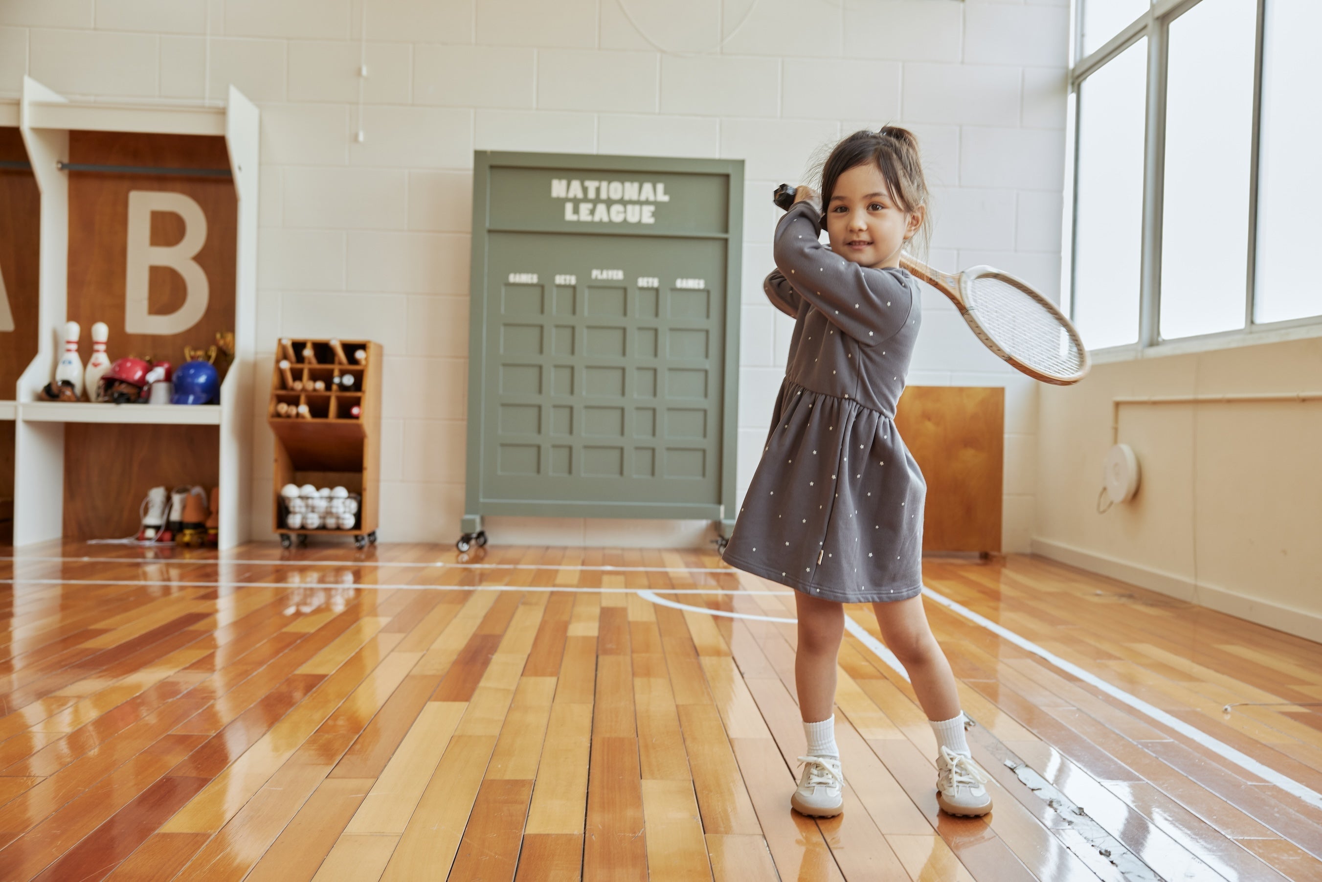 Child in a gray dress holding a badminton racket in an indoor setting with wooden floor and large window.