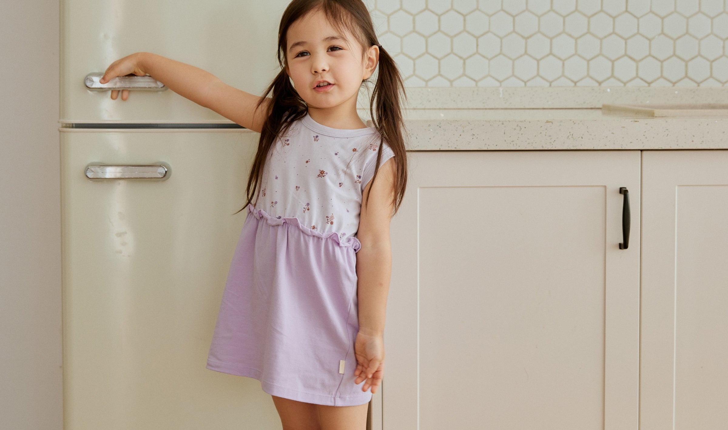 toddler girl wearing a purple dress and standing beside the fridge