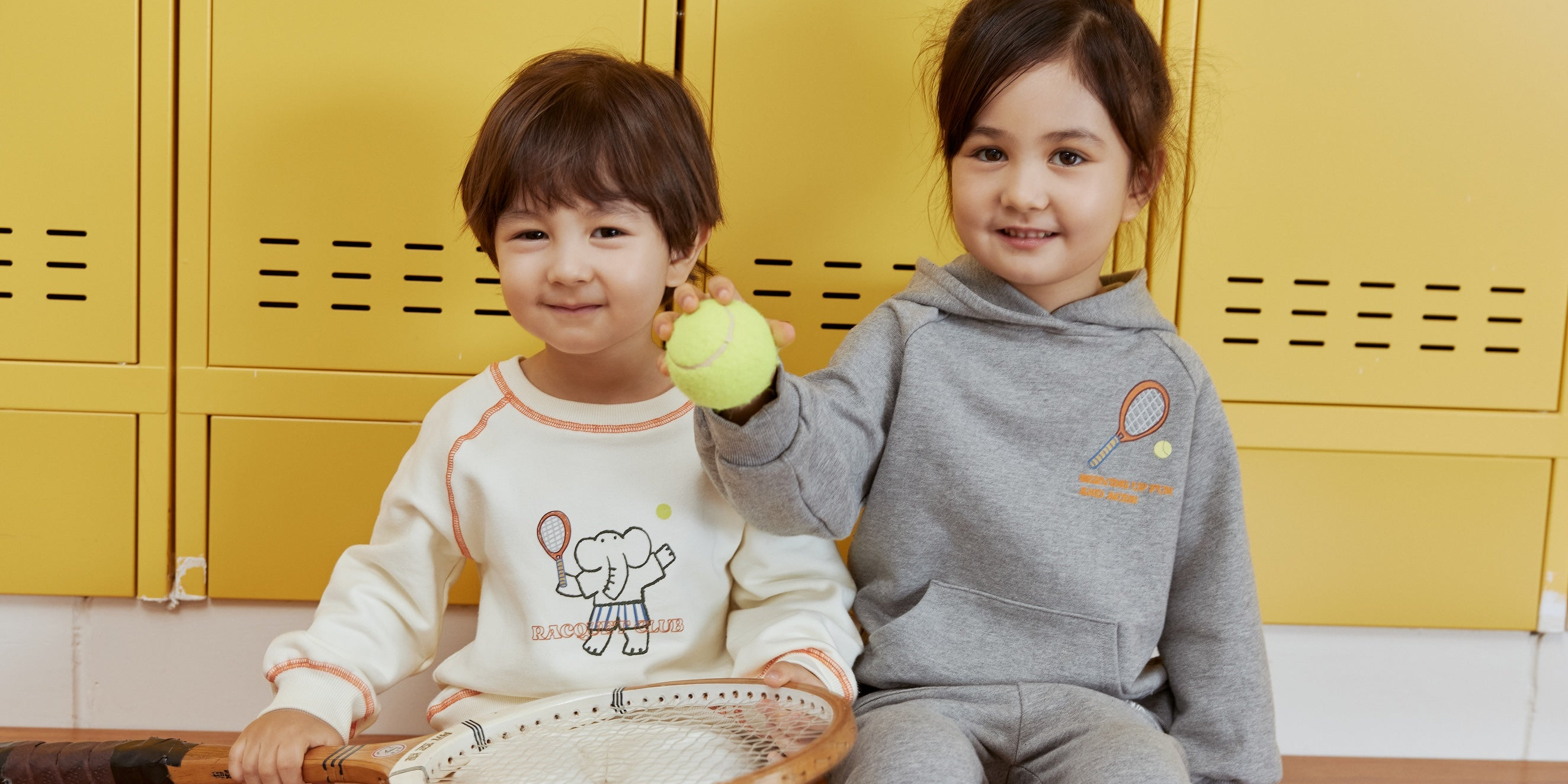 Two children sitting on a wooden floor with yellow lockers in the background.
