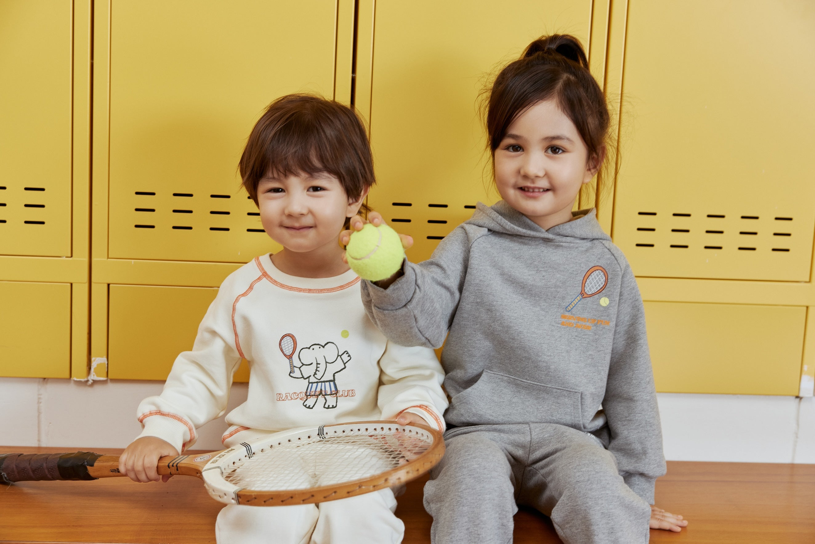Two children sitting on a wooden floor with yellow lockers in the background.