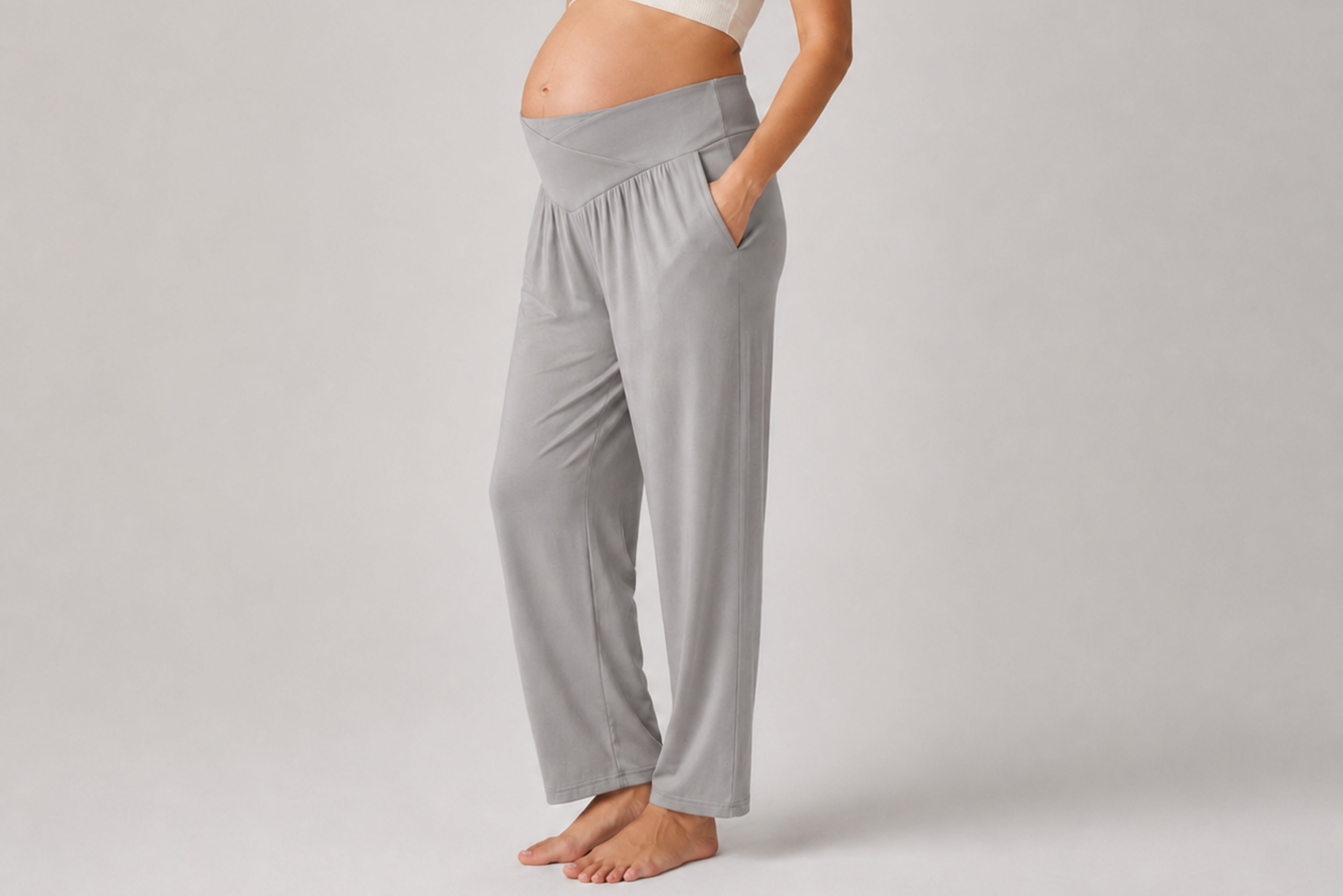 A pregnant model wearing light gray maternity lounge pants with side pockets, standing barefoot against a neutral light gray studio background.