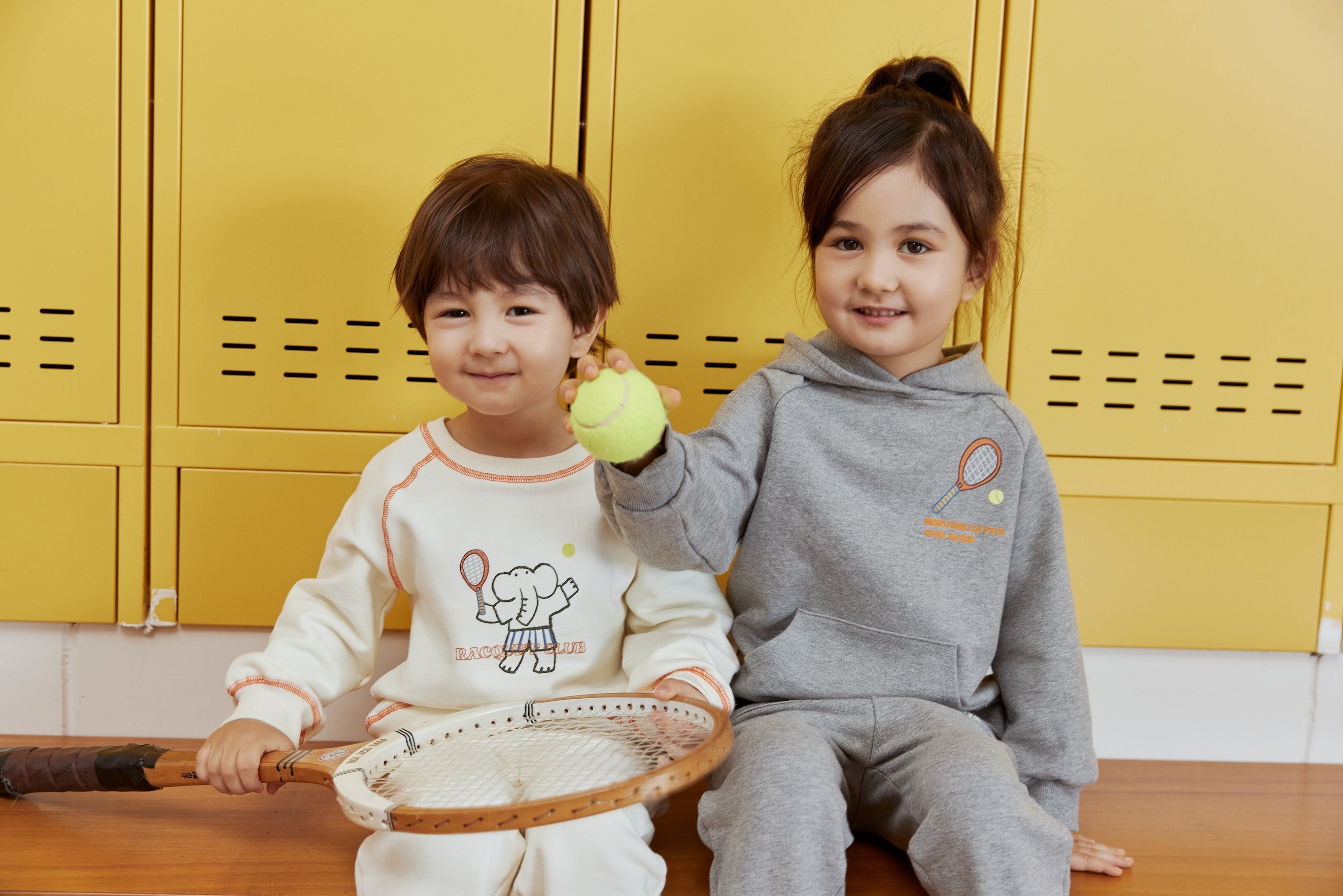 Two children sitting on a wooden floor with yellow lockers in the background.