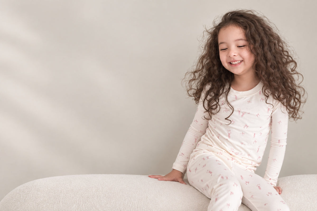 A young girl with long, curly brown hair sits smiling on a textured white cushion, wearing a matching long-sleeve pajama set with a delicate pink floral pattern against a soft beige background.