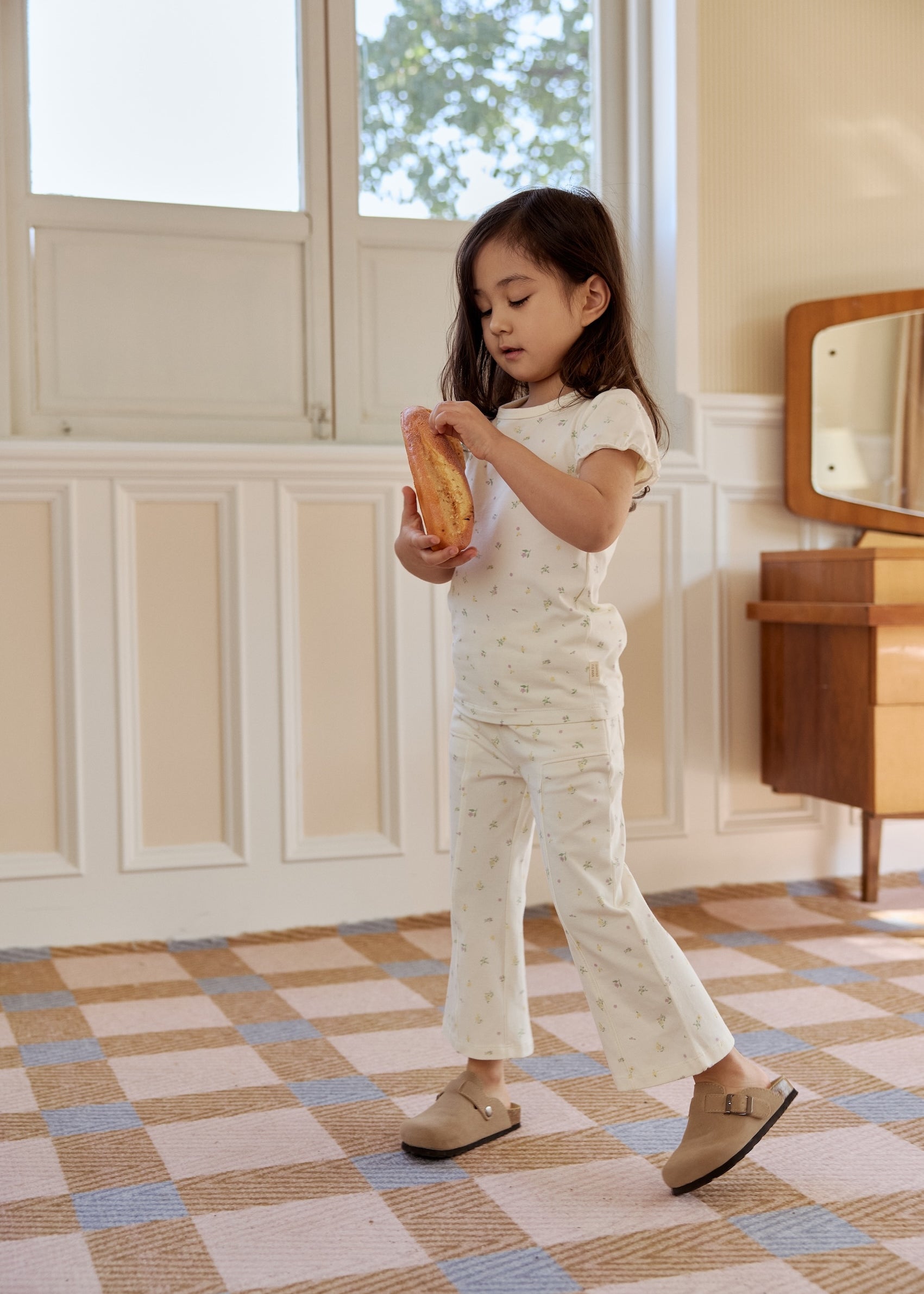  A little girl stands in a room with a white wall and a blue and white checkered floor while holding a bread
, smiling and looking around