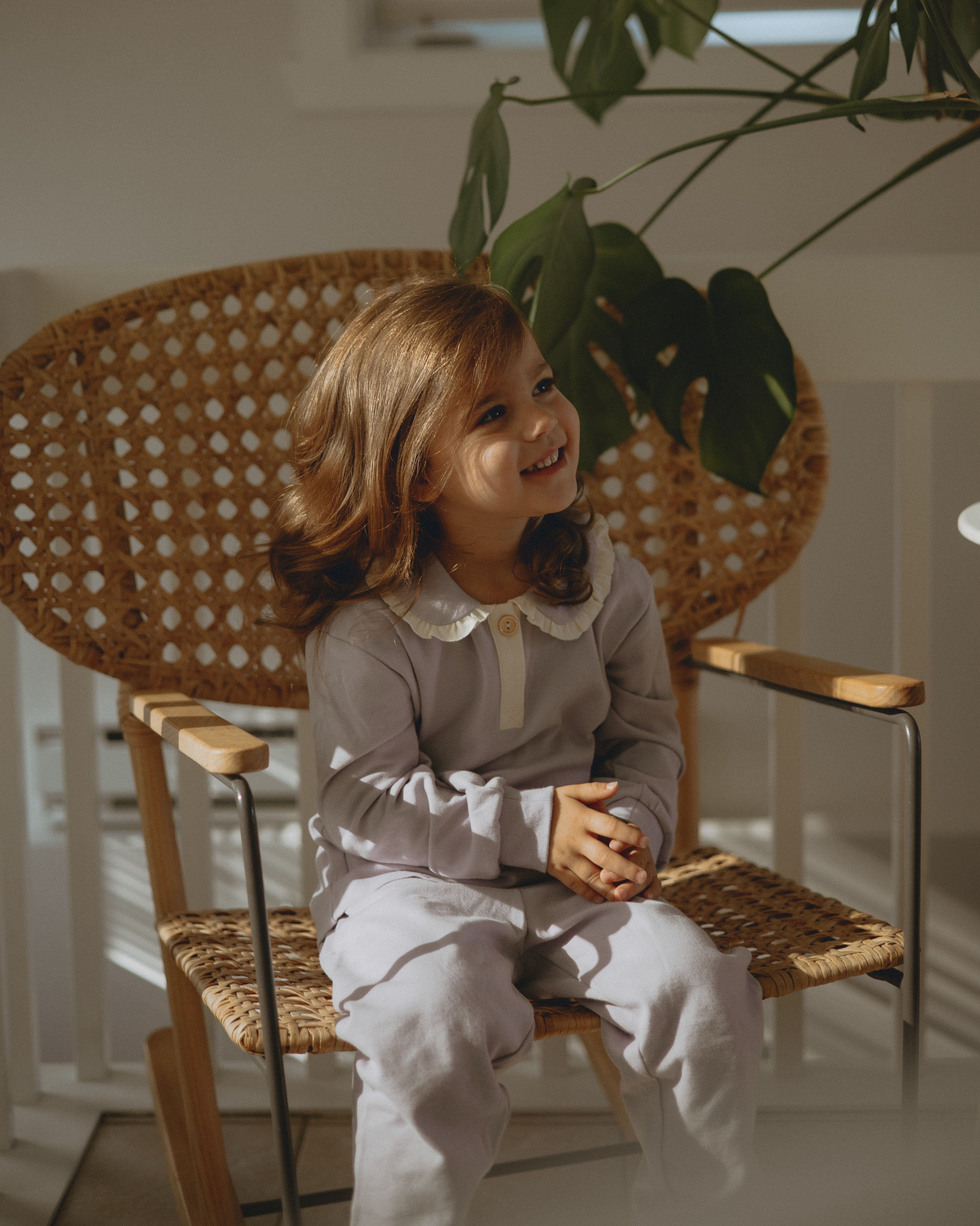 toddler girl is wearing norsu organic toddler polo shirt in lillac and sitting on the chair , smilling