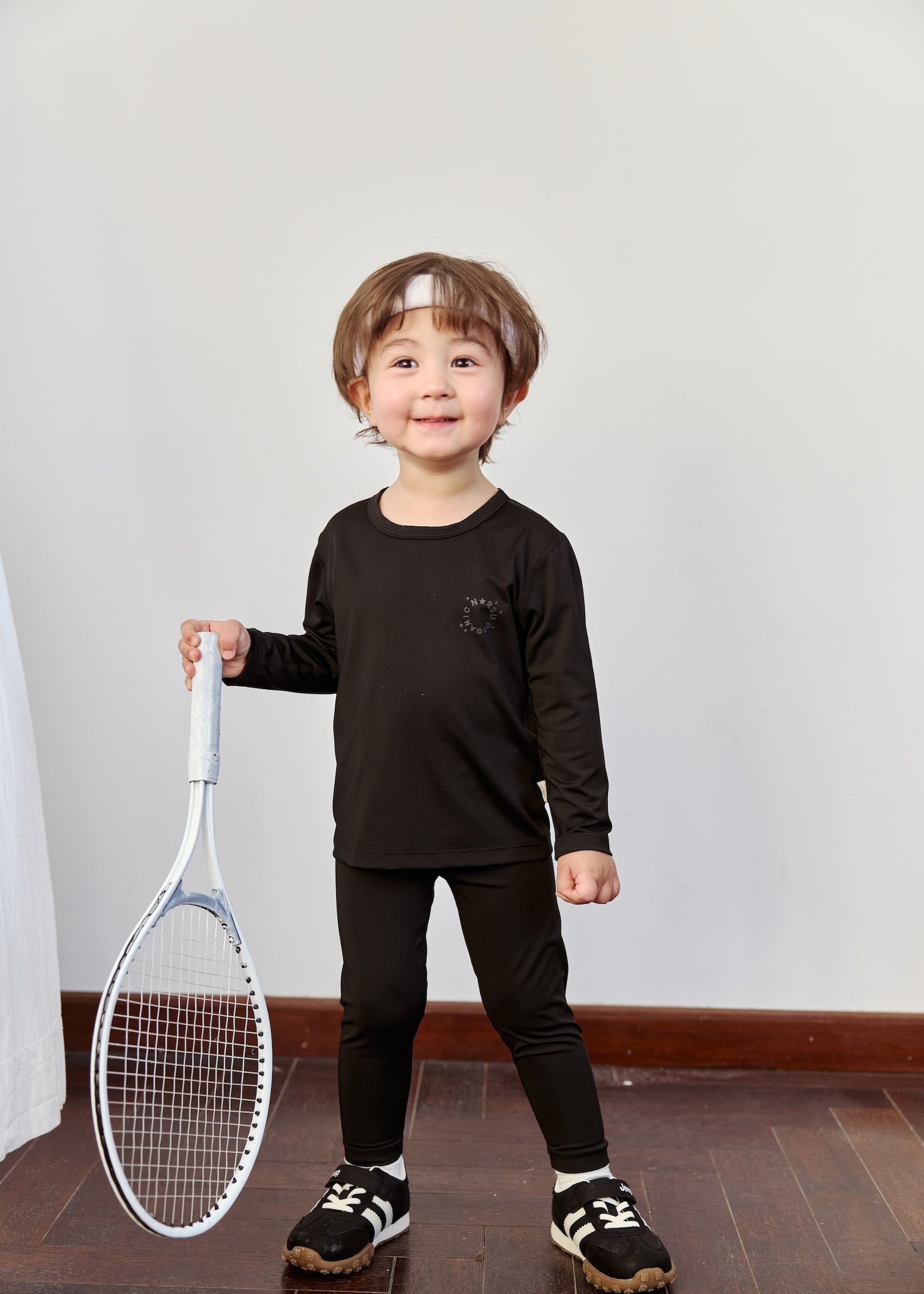 A young boy in a Toddler Dri-FIT long-sleeve shirt proudly holds a tennis racket, ready to play