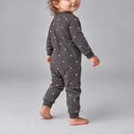 A toddler wearing a dark gray long-sleeve one-piece jumpsuit with a white star pattern, shown from the back as they walk against a light gray studio background.