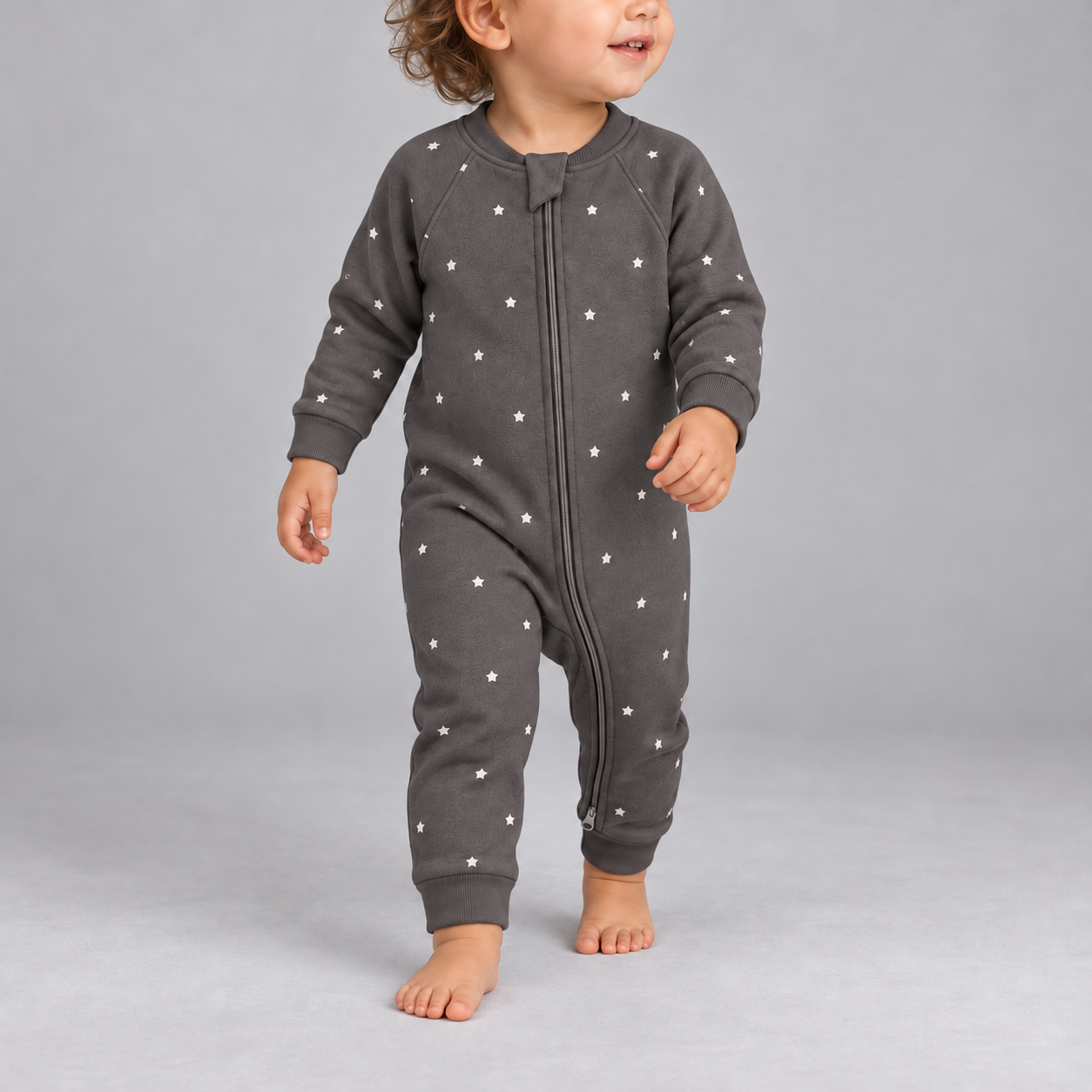 A baby wearing a dark gray long-sleeve footless sleeper with a white star pattern, walking barefoot against a solid gray studio background.