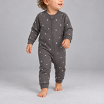 A baby wearing a dark gray long-sleeve footless sleeper with a white star pattern, walking barefoot against a solid gray studio background.