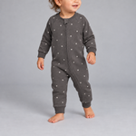 A toddler wearing a dark gray one-piece zip-up sleeper with a white star pattern, standing against a neutral gray studio background.