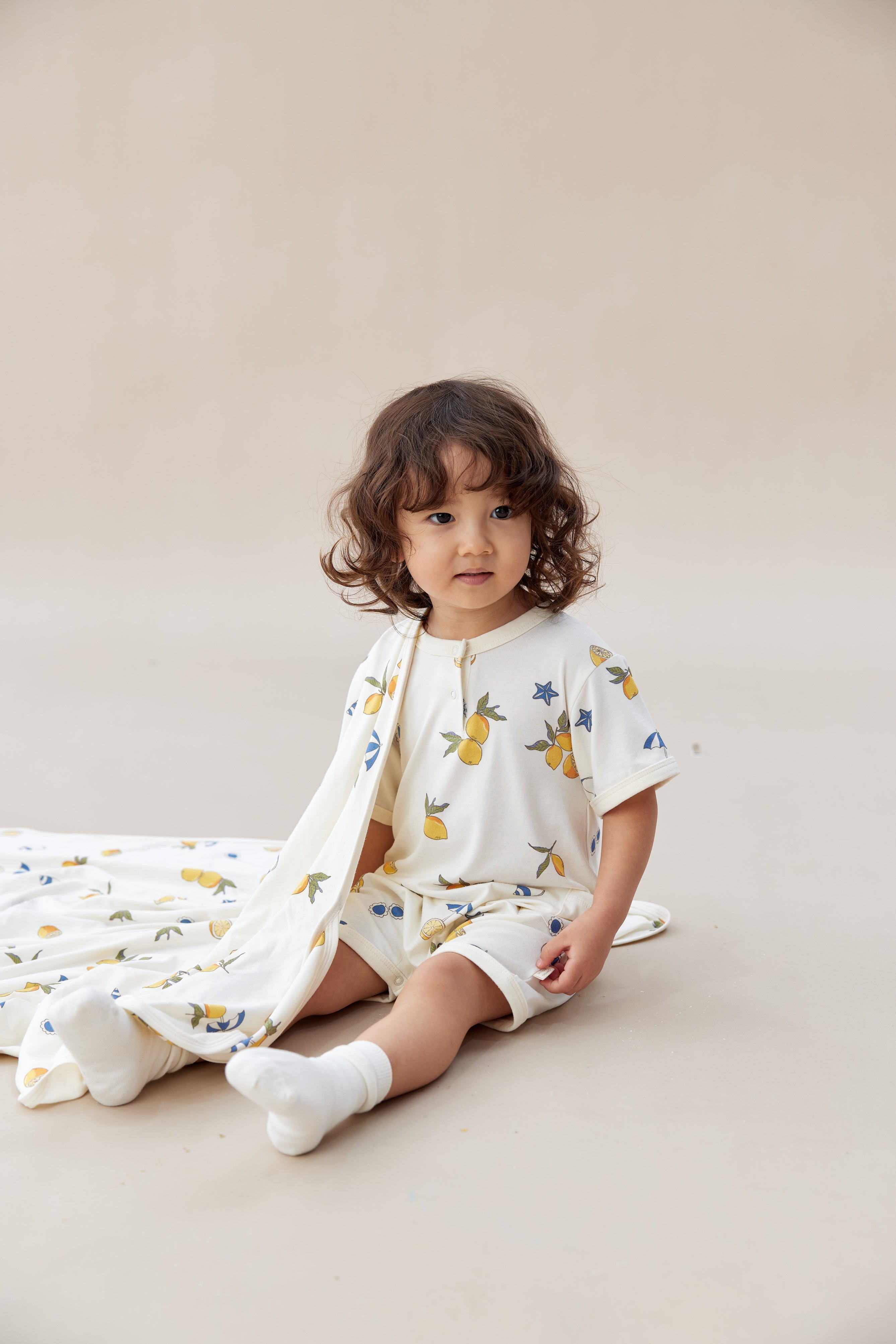 Child wearing a lemon romper with colorful patterns sitting on a beige surface.