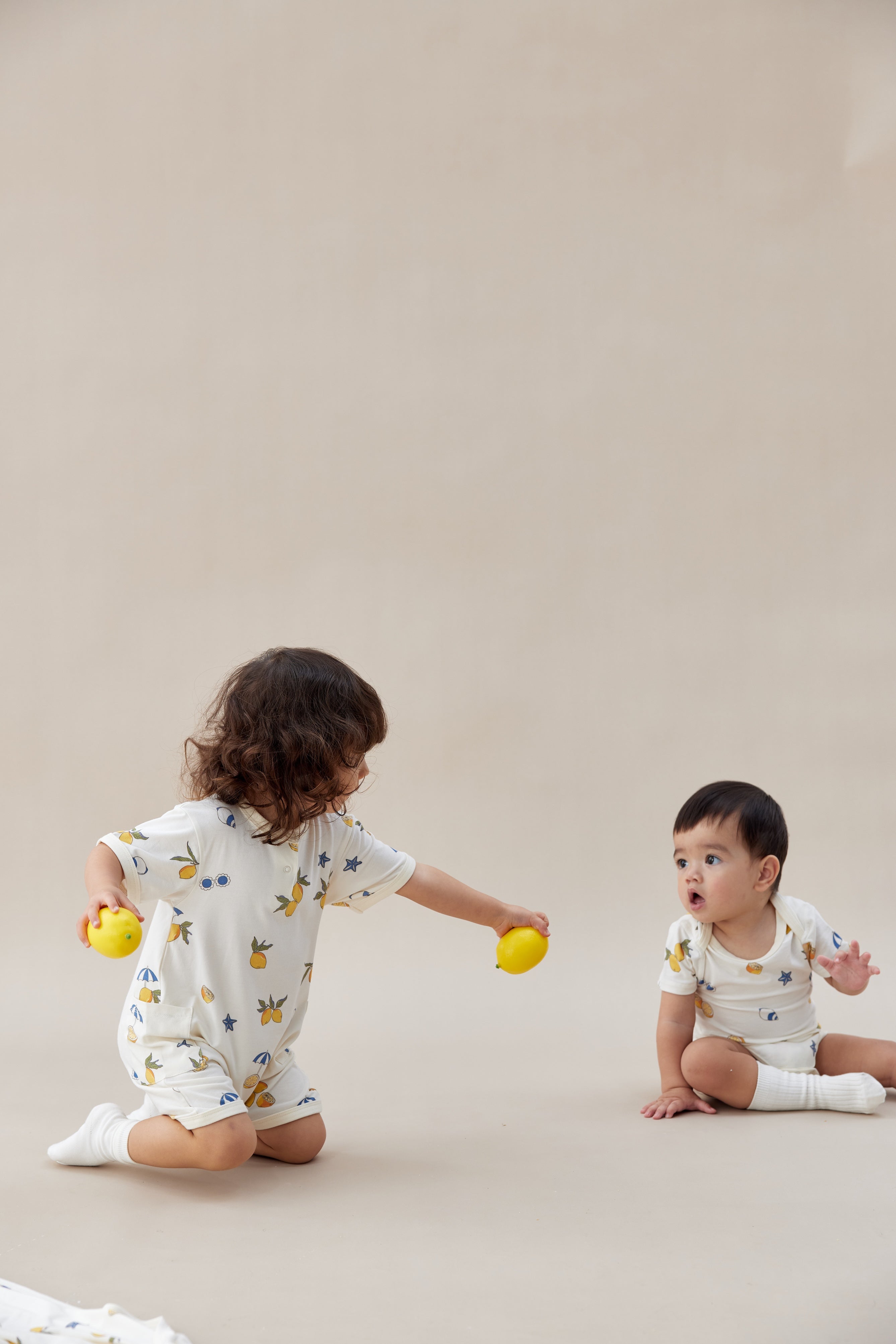 Two children in white onesies with colorful patterns playing on a beige surface.