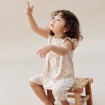 Child sitting on a small wooden stool wearing a sleeveless top and shorts with a pattern, pointing upwards.