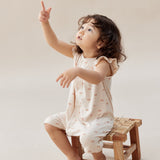 Child sitting on a small wooden stool wearing a sleeveless top and shorts with a pattern, pointing upwards.