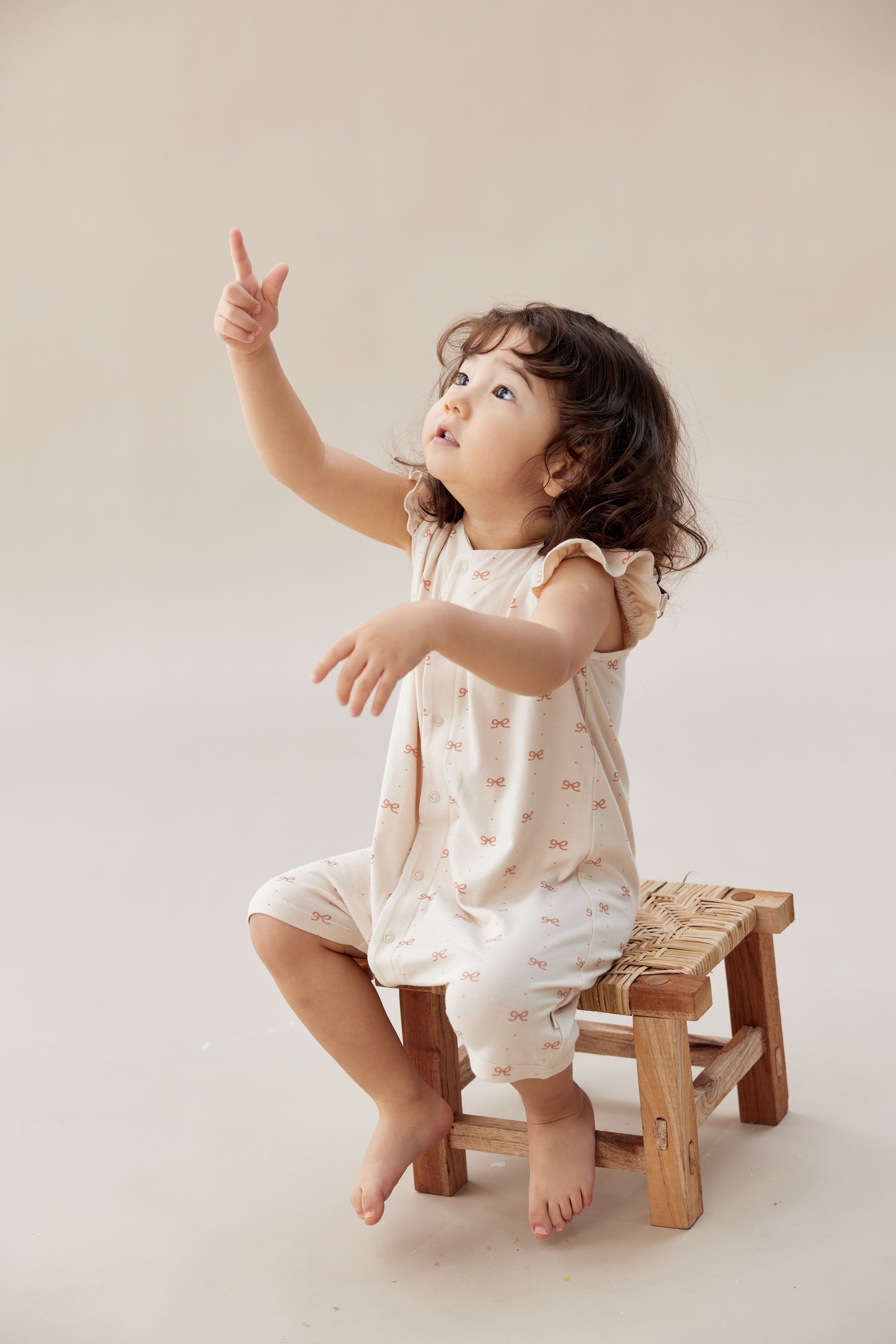 Child sitting on a small wooden stool wearing a sleeveless top and shorts with a pattern, pointing upwards.