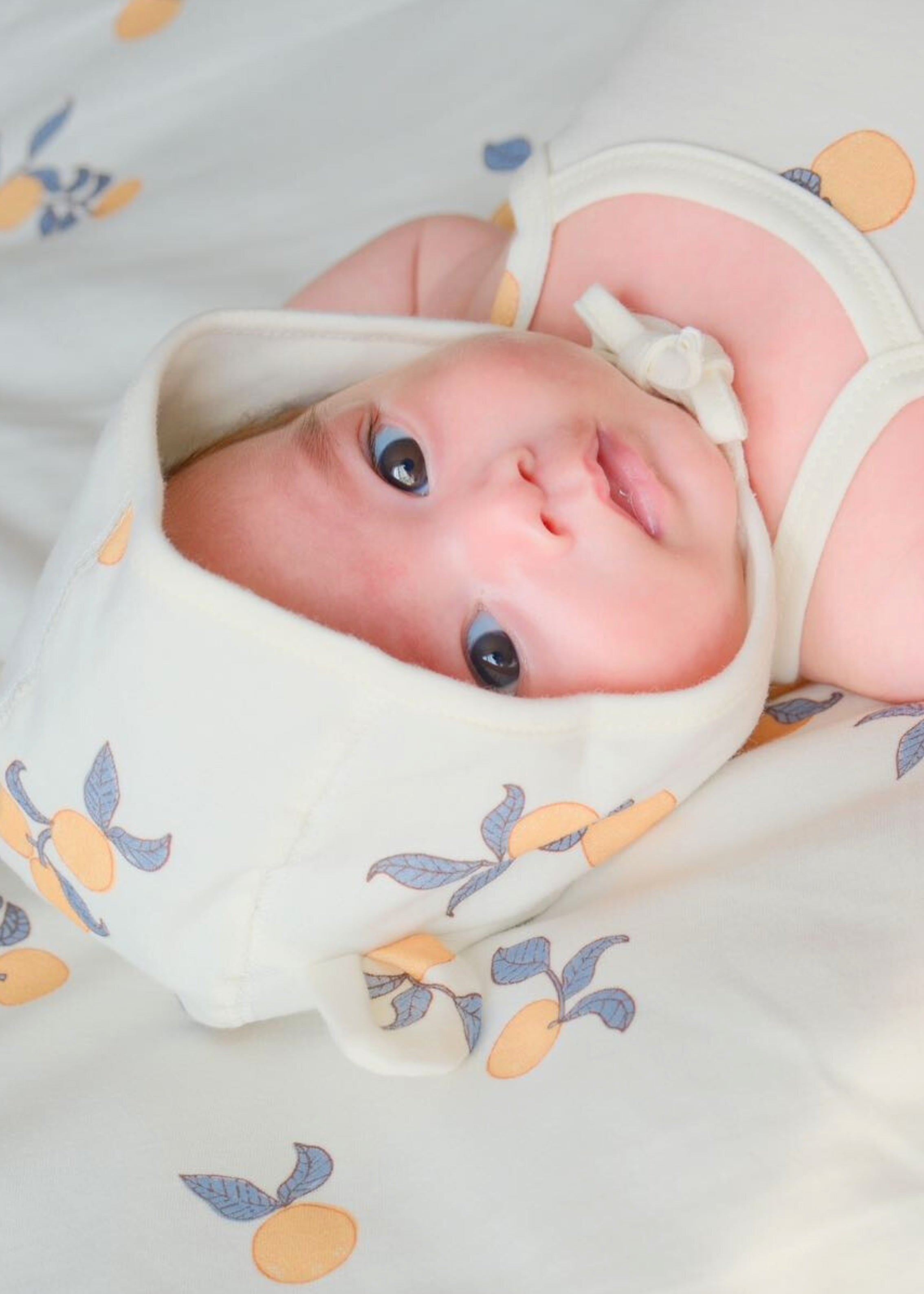 a baby in a orange print bed and wearing norsu organic baby bonnet hat in orange print and watching the camera