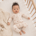 A baby lying on a white mattress in a wooden crib, wearing a cream-colored zip-up footed sleeper with small brown heart prints. A beige cable-knit blanket is draped on the left side of the crib.