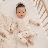 A baby lying on a white mattress in a wooden crib, wearing a cream-colored zip-up footed sleeper with small brown heart prints. A beige cable-knit blanket is draped on the left side of the crib.