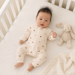 A baby lying on a white crib mattress wearing a cream-colored zip-up footless sleeper with small brown heart prints. A plush bunny toy and a white knit blanket are beside the baby.