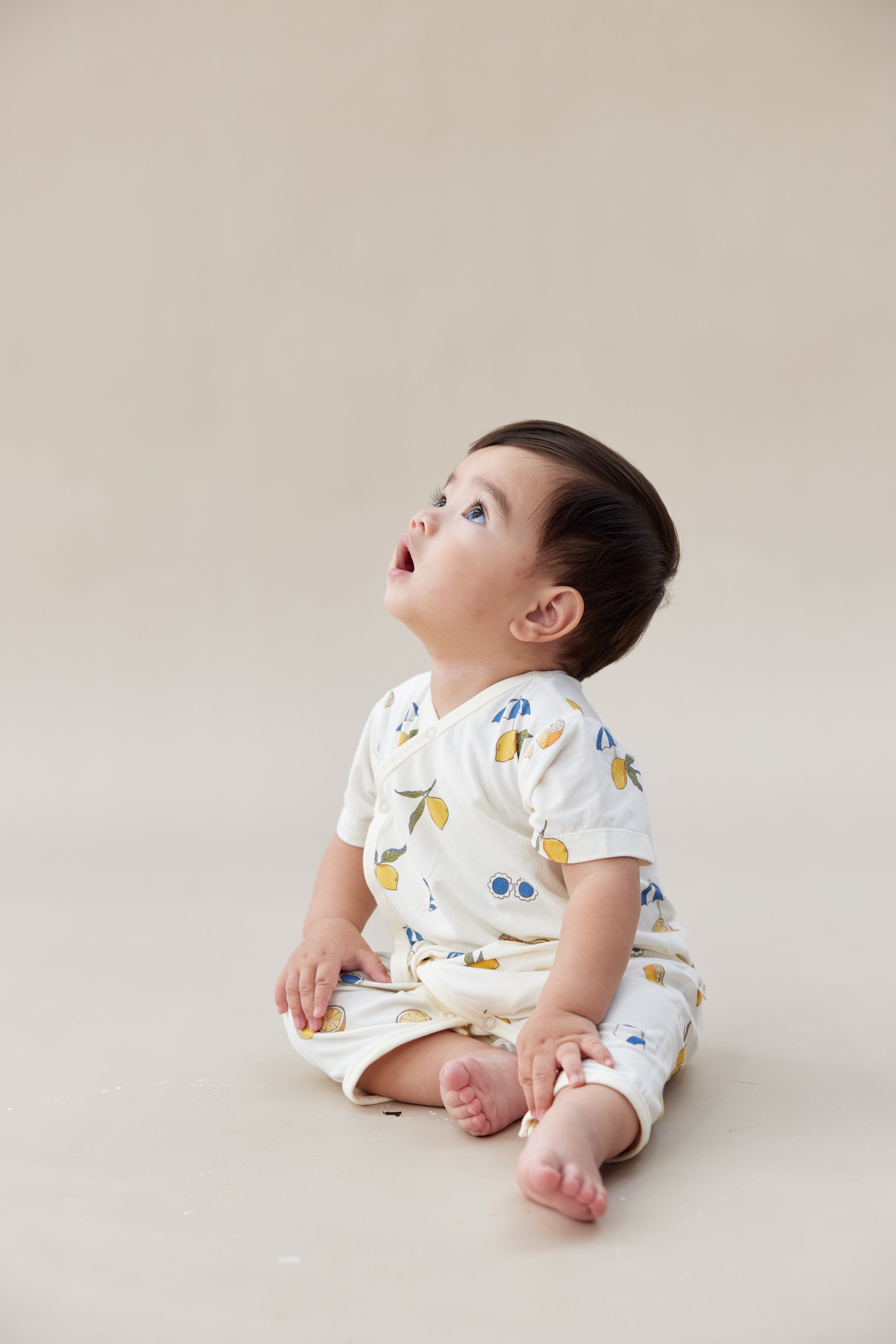 Baby sitting on a beige surface wearing a white outfit with colorful patterns.