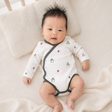 A baby lying on a cream-colored blanket next to a textured pillow, wearing a white long-sleeve bodysuit with dark gray trim and small penguin prints.