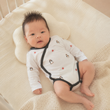 A baby lying on a cream-colored textured blanket in a crib, wearing a white long-sleeve bodysuit with black trim and small penguin prints.