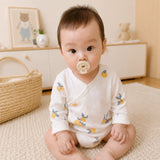 A baby sitting on a cream-colored rug in a warm, neutral-toned nursery, wearing a white long-sleeve wrap bodysuit with a yellow lemon and blue leaf pattern. The baby has a pacifier in their mouth.