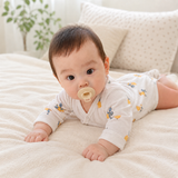 A baby lying on a cream-colored blanket, wearing a white long-sleeve onesie with small yellow and blue butterfly prints. The baby has a beige pacifier in their mouth, with pillows and a soft, blurred room background.