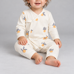 A baby sitting and wearing a cream-colored long-sleeve footless romper with a printed pattern of orange fruits and blue-gray birds, photographed against a neutral gray background.