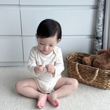 A baby sitting on a textured cream rug in front of white paneled walls, wearing a cream long-sleeve bodysuit with a small pink floral pattern. A woven basket with a brown plush toy is on the right.