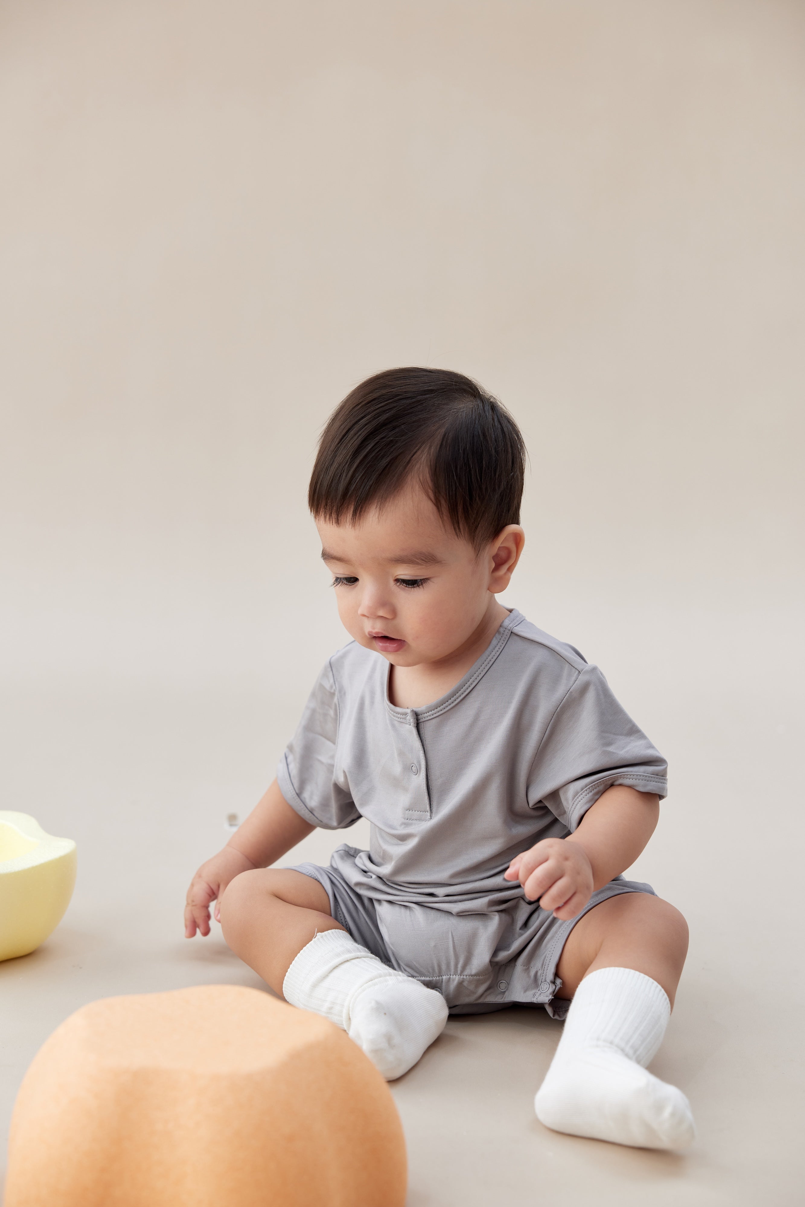 Baby in a gray outfit sitting on a beige surface with a white background