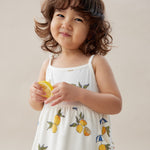 Child wearing a white dress with lemon prints, holding a lemon against a plain background