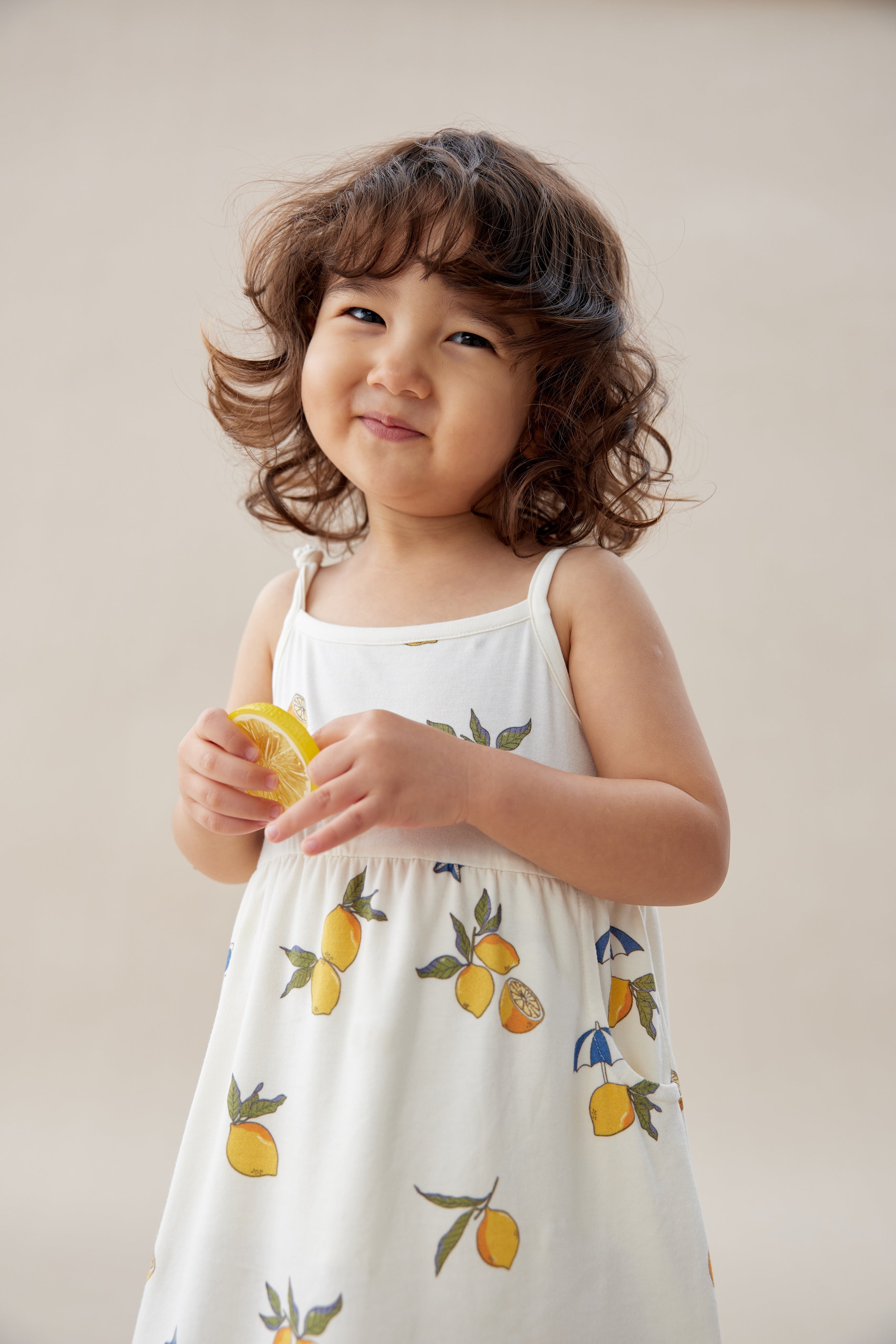 Child wearing a white dress with lemon prints, holding a lemon against a plain background