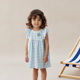 Young girl wearing a blue and white striped dress standing next to a deck chair on a beige background