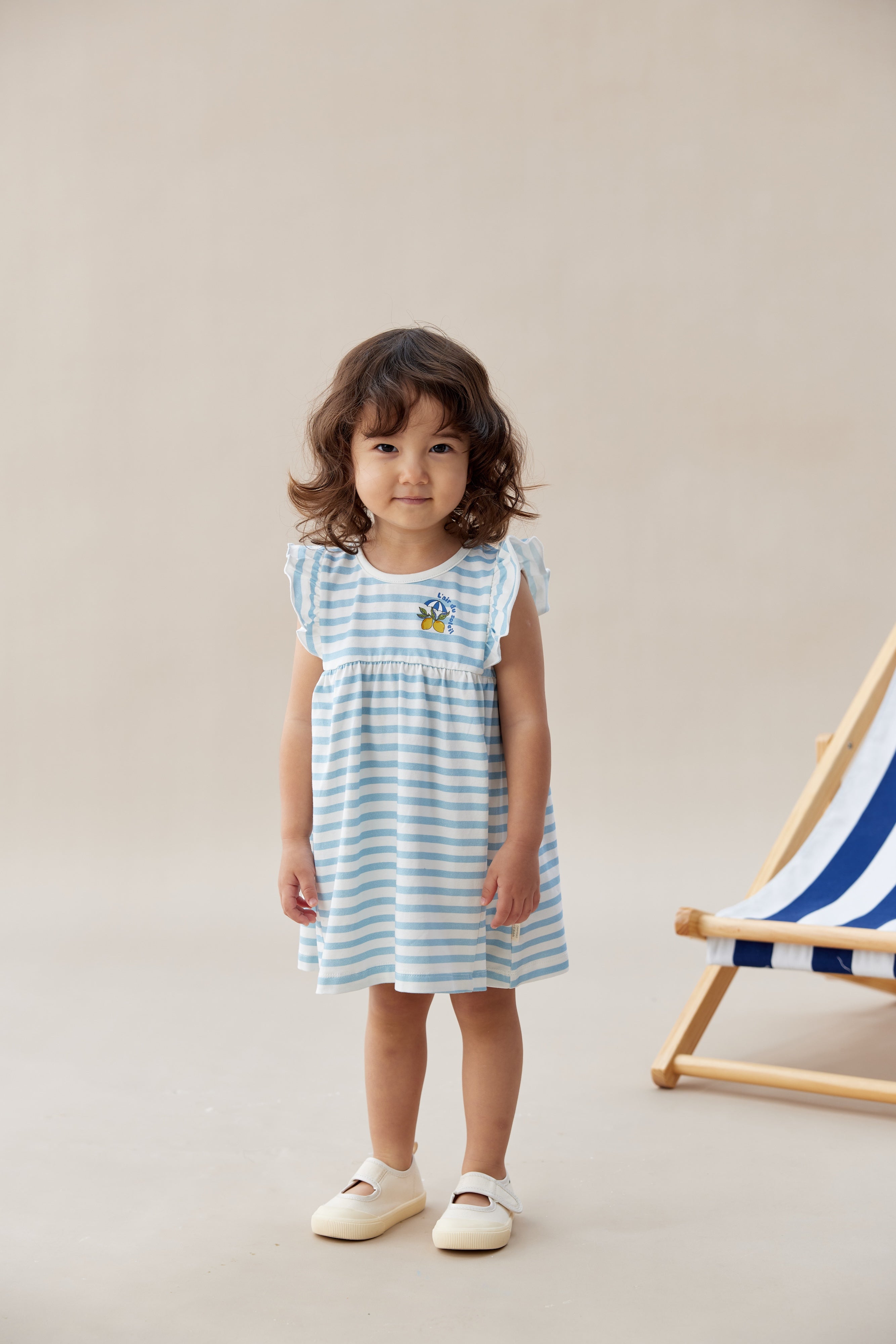 Young girl wearing a blue and white striped dress standing next to a deck chair on a beige background