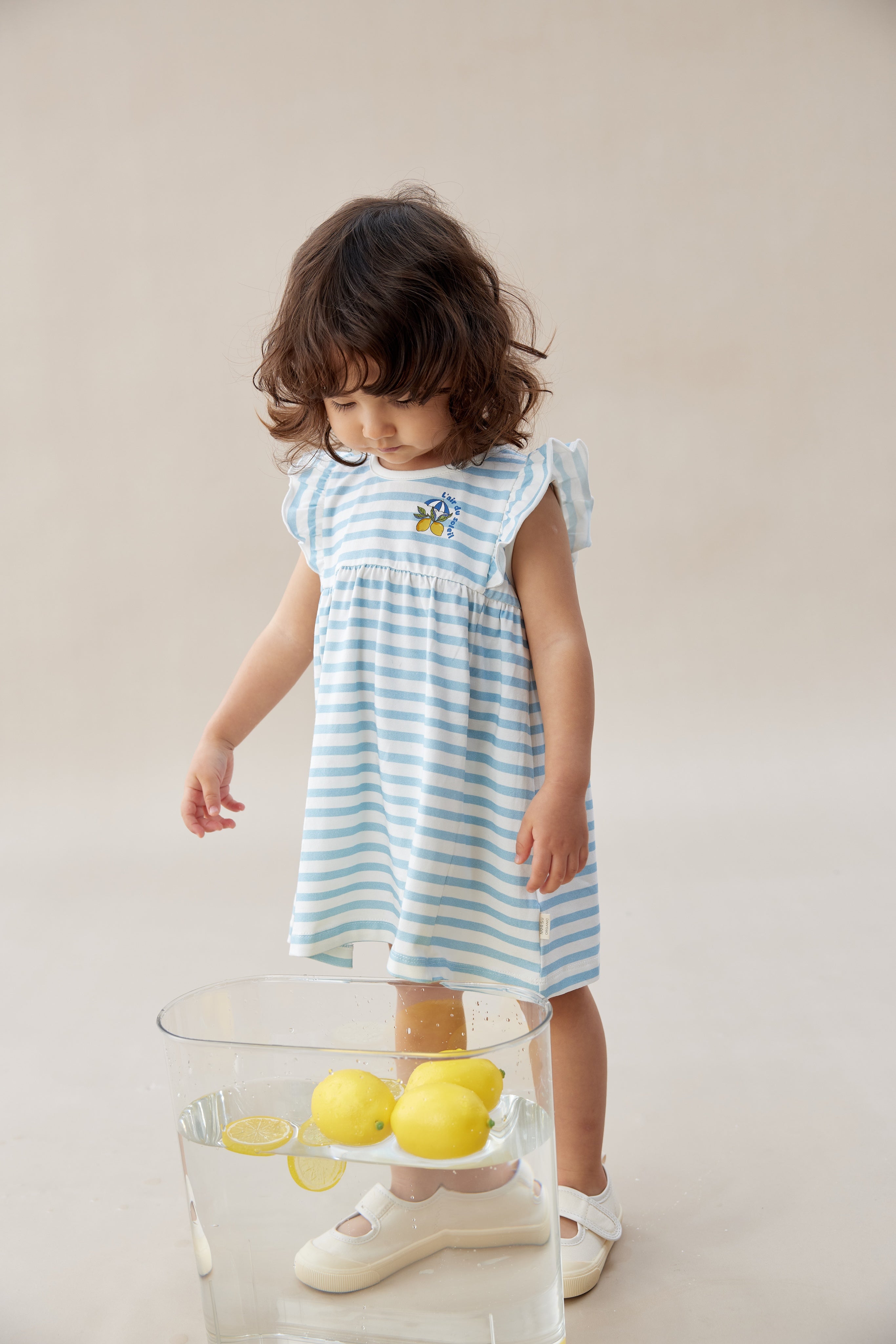 Young girl in a blue and white striped dress standing next to a glass table with lemons on a plain background
