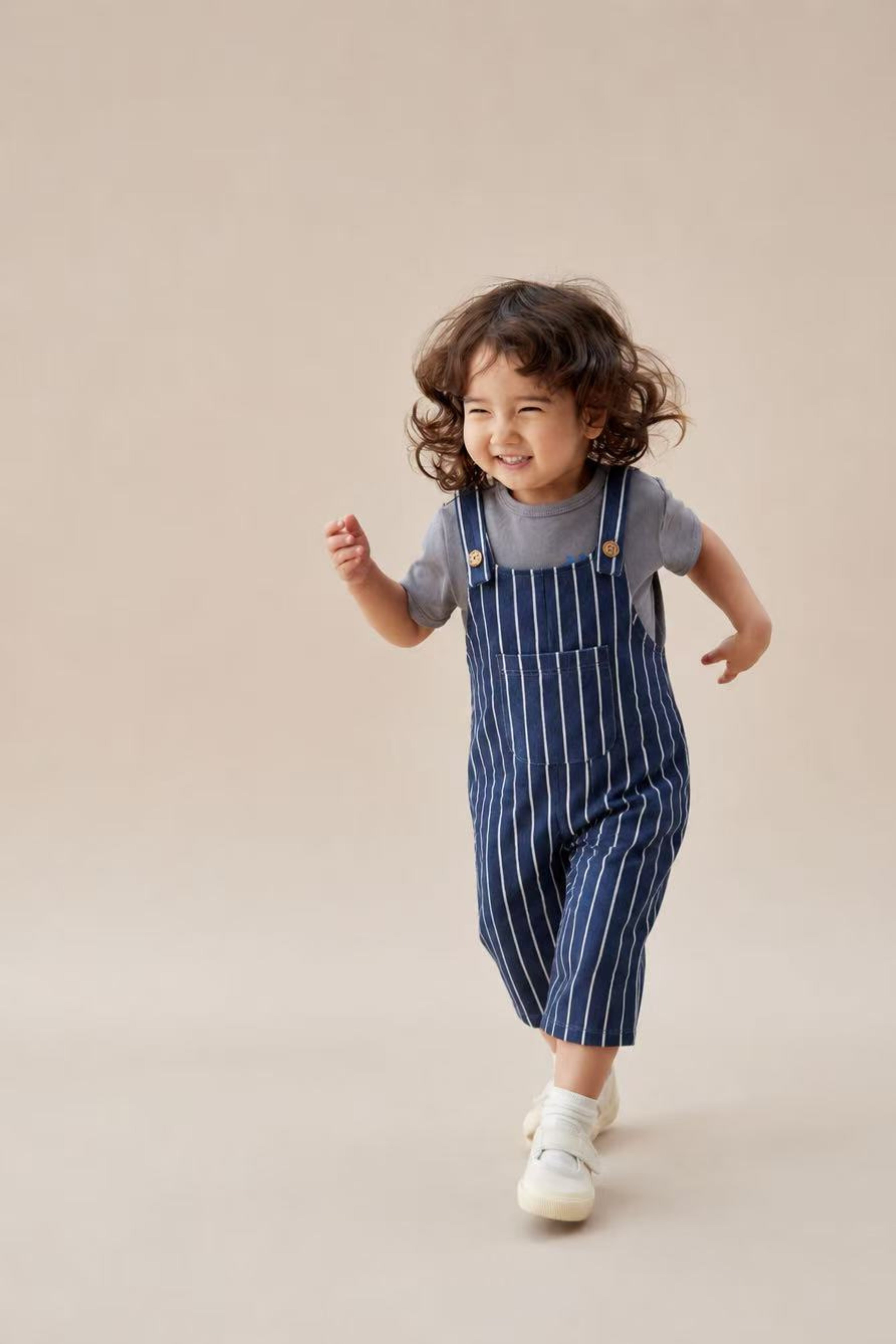 A toddler girl with curly brown hair is captured mid-run, wearing a navy blue and white vertically striped overall jumpsuit over a grey t-shirt and white sneakers, set against a solid warm beige studio background.