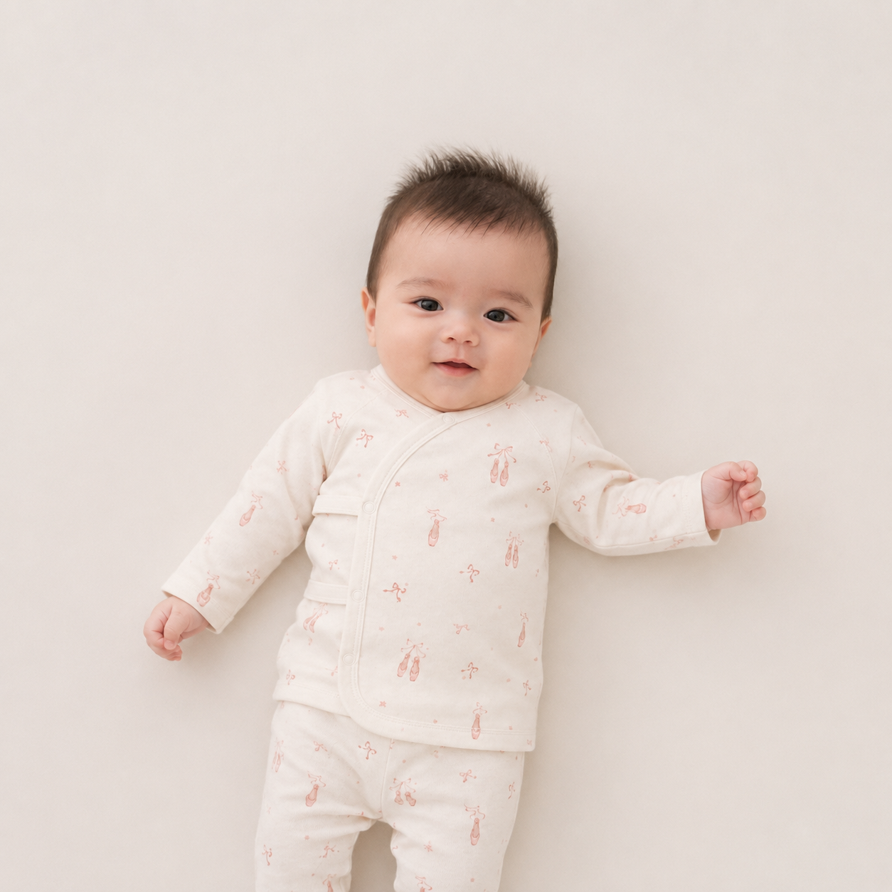 A baby lying on their back wearing a cream-colored long-sleeve pajama set with a small pink bunny pattern, photographed against a solid light beige background.