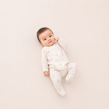 A baby lying on their back wearing a white long-sleeve footed sleeper with a tiny pink pattern, photographed on a solid light beige background.