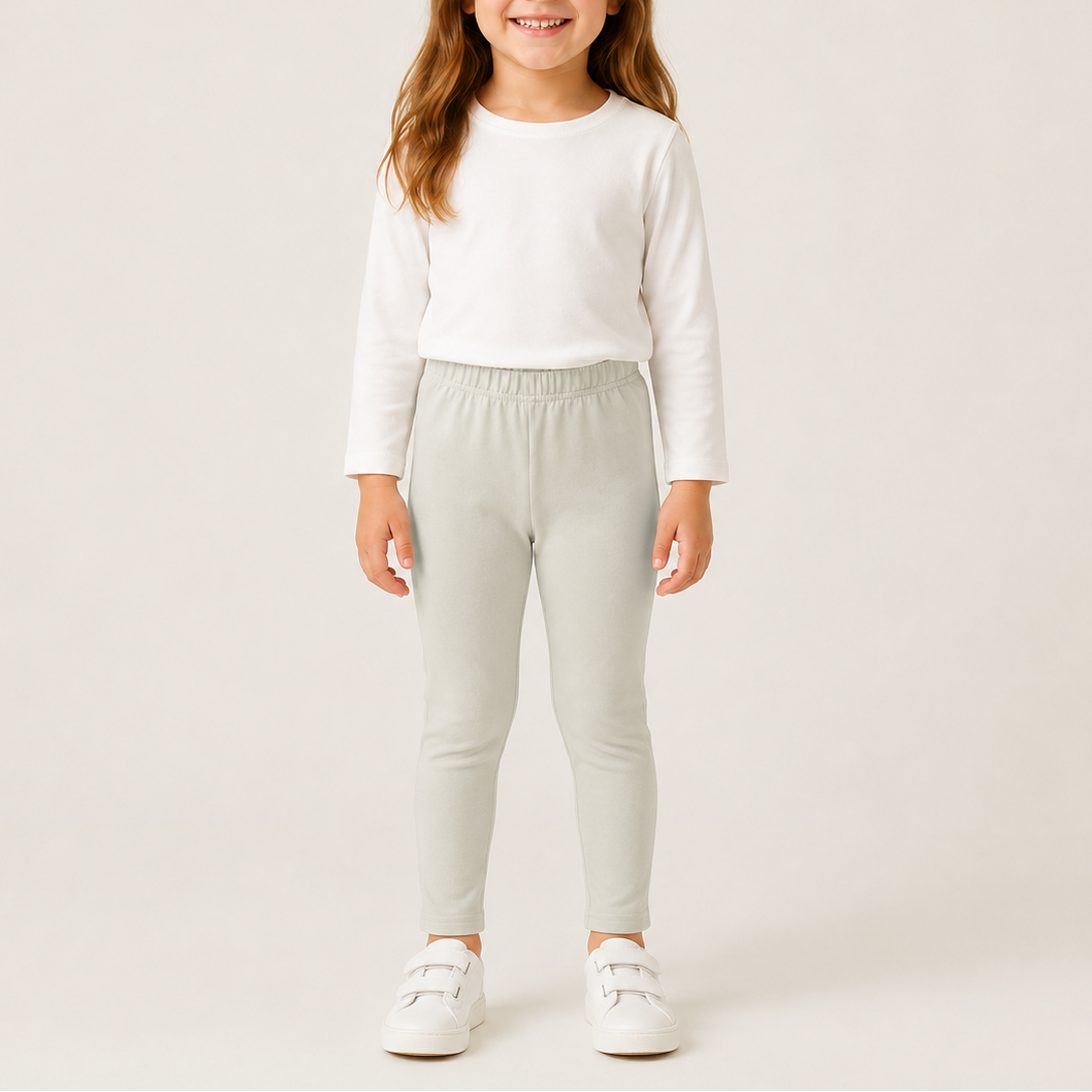 A young girl modeling light sage green slim-fit pants, paired with a white long-sleeve top and white sneakers, against a plain light beige studio background.