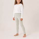 A young girl modeling a white long-sleeve top and light sage green leggings against a plain, warm off-white studio background.