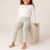 A young girl with long brown hair sits barefoot on a light beige block, wearing a white long-sleeve crewneck shirt and light sage green leggings against a warm off-white studio background.