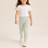A young girl walking forward in a white short-sleeve t-shirt and light sage green jogger pants, paired with white sneakers, against a solid light beige studio background.