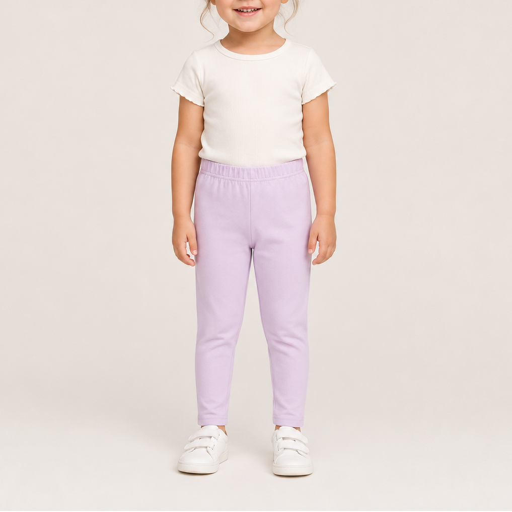A young child standing against a plain light beige studio background, wearing a white short-sleeve top, light lavender pants, and white sneakers.