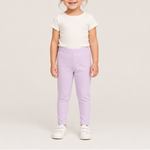 A young child standing against a plain light beige studio background, wearing a white short-sleeve top, light lavender pants, and white sneakers.
