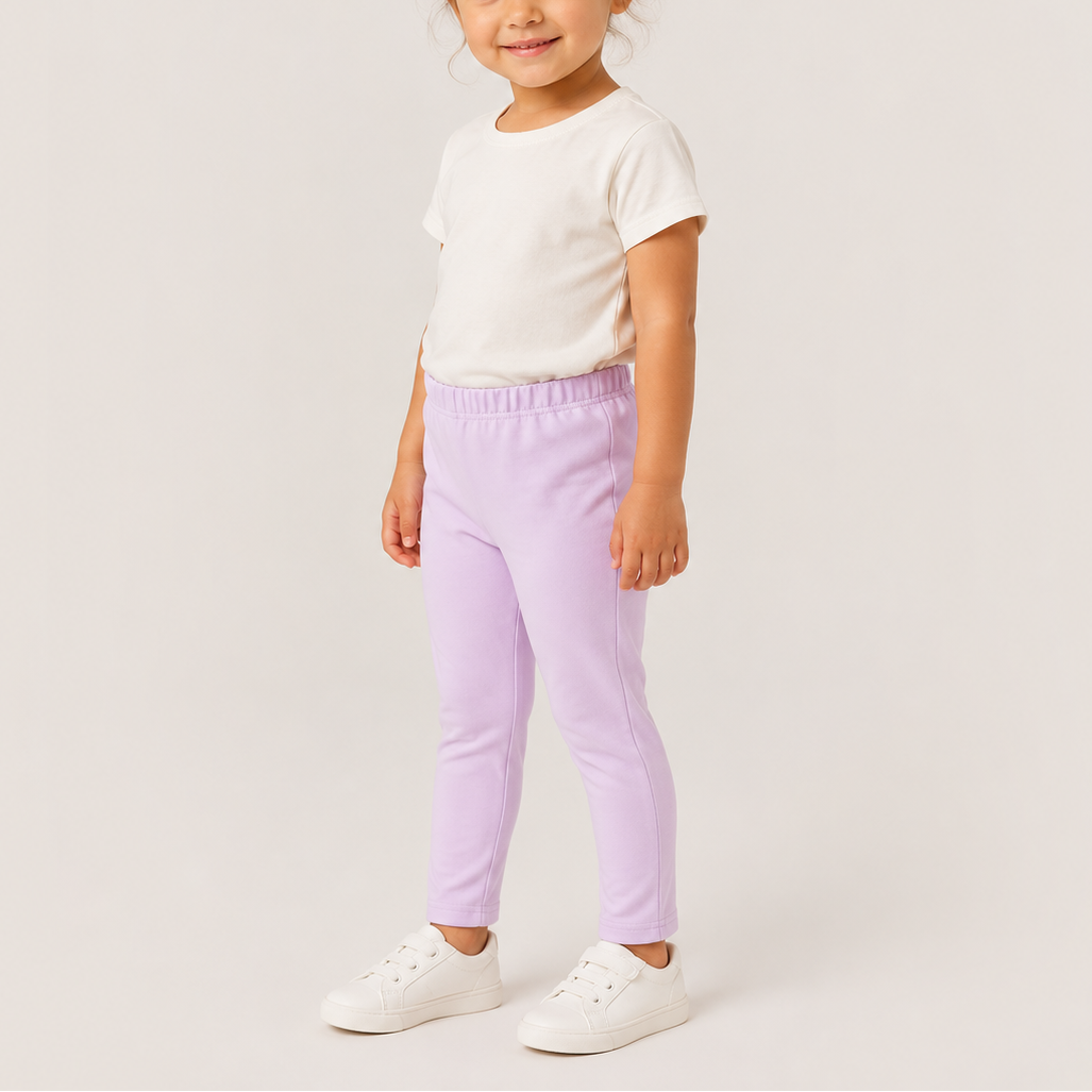 A young girl with her hair in a bun stands against a light gray studio background, wearing a white short-sleeve t-shirt, light purple elastic-waist pants, and white sneakers.