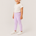 A young girl with her hair in a bun stands against a light gray studio background, wearing a white short-sleeve t-shirt, light purple elastic-waist pants, and white sneakers.