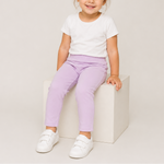 A young child wearing a white t-shirt and light purple cropped pants, sitting on a white block against a light beige studio background.